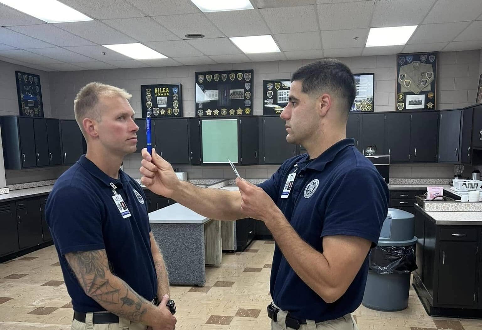 Two men in navy uniforms stand in a room; one holds a pen and a notepad, performing an eye-tracking test on the other, who looks straight ahead. The room has cabinets, counters, and law enforcement insignias on the wall.