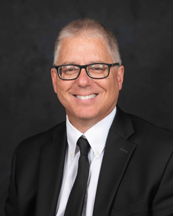 A middle-aged man with short gray hair and glasses, wearing a black suit, white shirt, and black tie, smiling at the camera against a dark background.