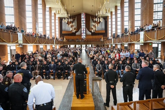 A large crowd, including many police officers in uniform, gathers inside a spacious church with high ceilings and tall windows for a ceremony or event. The focus is on a speaker addressing the audience from the front.