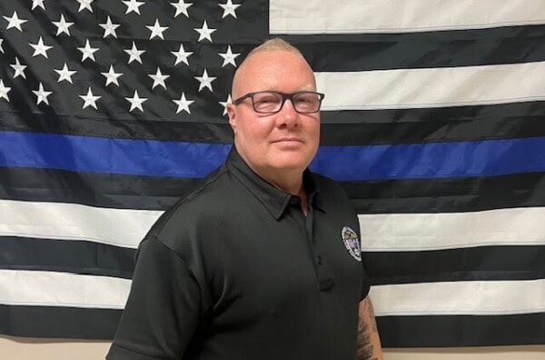 A man with short light hair, glasses, and a black collared shirt stands in front of a black-and-white American flag featuring a thin blue line.