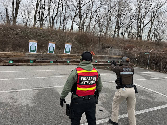 Two people stand at a shooting range. One wears a vest labeled FIREARMS INSTRUCTOR, while the other, labeled SHERIFF, aims a handgun at a paper target. Targets are set against a berm with leafless trees.