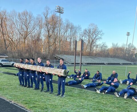 A group of people in matching athletic gear lift a large log together on a grassy field, while others lie on exercise mats nearby. Bleachers and leafless trees are visible in the background.