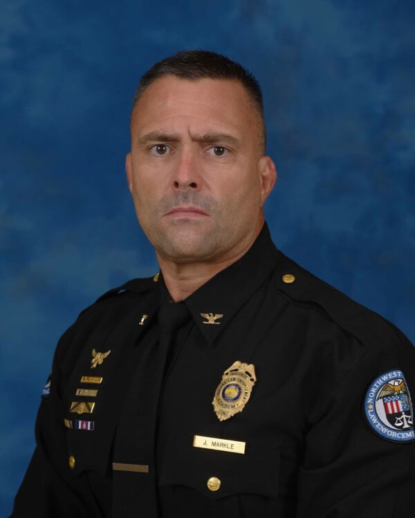 A man in a police uniform with badges and insignia poses against a blue background, looking directly at the camera with a serious expression.
